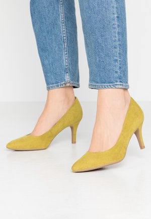 Geel suède hoge hak schoenen met een spitse neus en een slanke hak, gedragen met spijkerbroek op enkelhoogte. Gladde textuur en eenvoudig ontwerp.