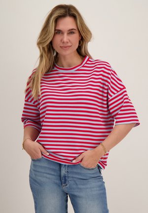 Vrouw met lang blond haar die een rood en lichtblauw gestreept shirt en spijkerbroek draagt, staand met handen in de zakken tegen een effen achtergrond.