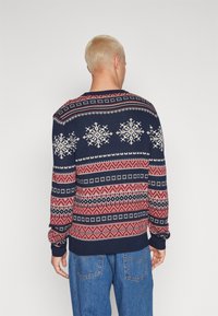 Maglione lavorato a maglia blu navy con motivo a fiocchi di neve bianchi ripetuto, accenti geometrici rossi e bianchi, polsini a coste e un classico scollo rotondo.