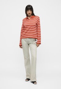 Femme portant un polo à manches longues rayé rouge et blanc, un pantalon beige à jambes larges et des sandales noires ouvertes aux orteils, debout sur un fond blanc.