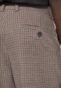 Spodnie w wzór houndstooth w odcieniach brązu i beżu, o dopasowanym kroju, z jedną tylnią kieszenią i czarnym guzikiem jako akcentem.