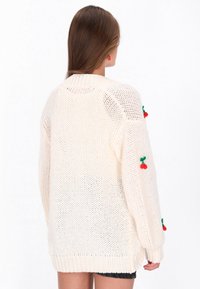 Sweter kardigan w kolorze kremowym o luźnym kroju, z zielonymi i czerwonymi akcentami w kształcie wiśni na rękawach oraz ściągaczami u dołu i na mankietach.