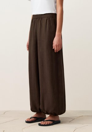 Person trägt lockere braune Hose mit elastischen Bündchen und Bund, kombiniert mit schwarzen Flip-Flops, steht auf gefliestem Boden.