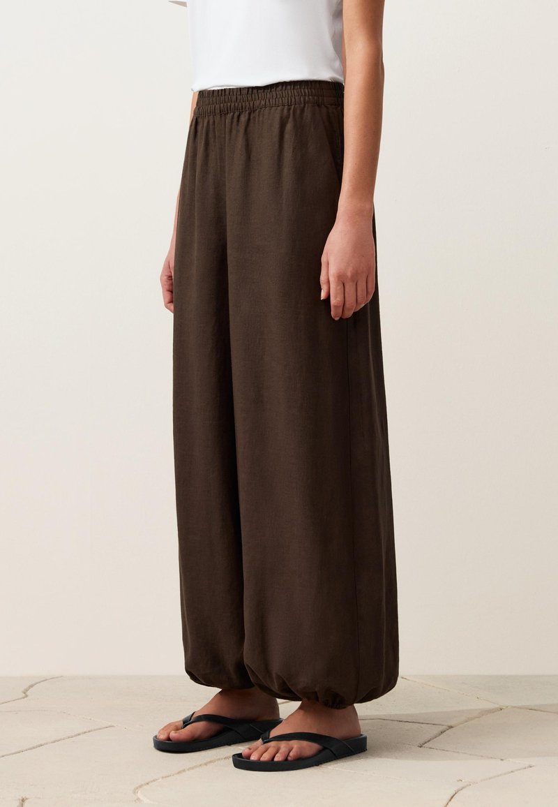 Personne portant un pantalon marron ample avec des poignets et une taille élastiques, associé à des tongs noires, debout sur un sol carrelé.