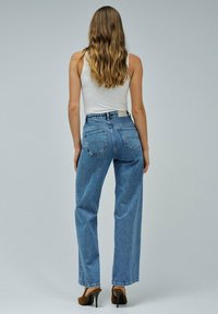 Mujer con cabello largo y ondulado, vistiendo una blusa blanca sin mangas, jeans de tiro alto de pierna recta azules y zapatos de tacón marrones, de espaldas.