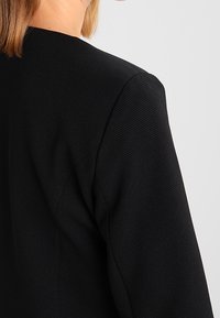 Blazer noir en tissu texturé, avec un col montant et des épaules structurées. Le dos présente des coutures invisibles pour un look épuré.