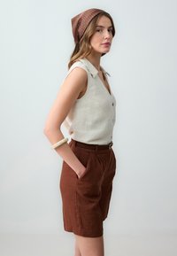 Camicia senza maniche in lino di un leggero beige con colletto, abbinata a pantaloni corti in lino marroni e un foulard a fantasia. Accentata da un bracciale.