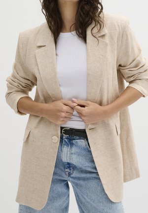 Mujer con blazer beige de mangas remangadas, top blanco acanalado, vaqueros azules y cinturón negro, ajustándose la parte frontal del blazer.
