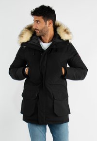 Parka noire avec une capuche bordée de fourrure, fermeture éclair et pression à l'avant, deux grandes poches avant et cordons ajustables à la taille.