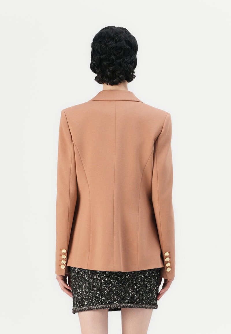 Różowa, dopasowana marynarka z poduszkami na ramionach, klapami i złotymi guzikami na mankietach, w zestawie z teksturowaną czarną mini spódnicą.