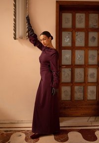 Ilgas purpurinis suknelė su sukrautomis rankovėmis ir surinkta juosmeniu. Juodi pirštinės akcentuoja dizainą. Medinės durys su raižytu floriniu stiklu tarnauja kaip fonas.