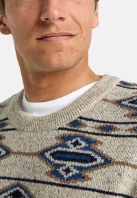 Suéter beige de punto con patrones geométricos en azul y marrón. Presenta un cuello redondo acanalado y una textura suave, adecuado para el uso casual.
