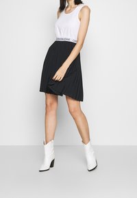 Vit ärmlös topp med en Calvin Klein midja, i kombination med en plisserad svart kjol och vita anklar stövlar. Neutral bakgrund.