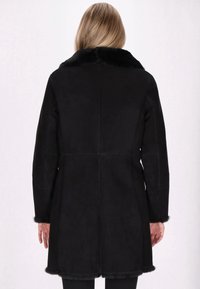 Cappotto in suede nero con colletto in pelliccia, maniche lunghe, taglio dritto e texture morbida. Presenta rifiniture pelose ai polsini e all'orlo.