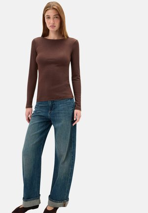 Giovane donna con lunghi capelli lisci che indossa un top marrone aderente a maniche lunghe, jeans blu a gamba larga con risvolti e scarpe scure, in piedi su sfondo bianco.