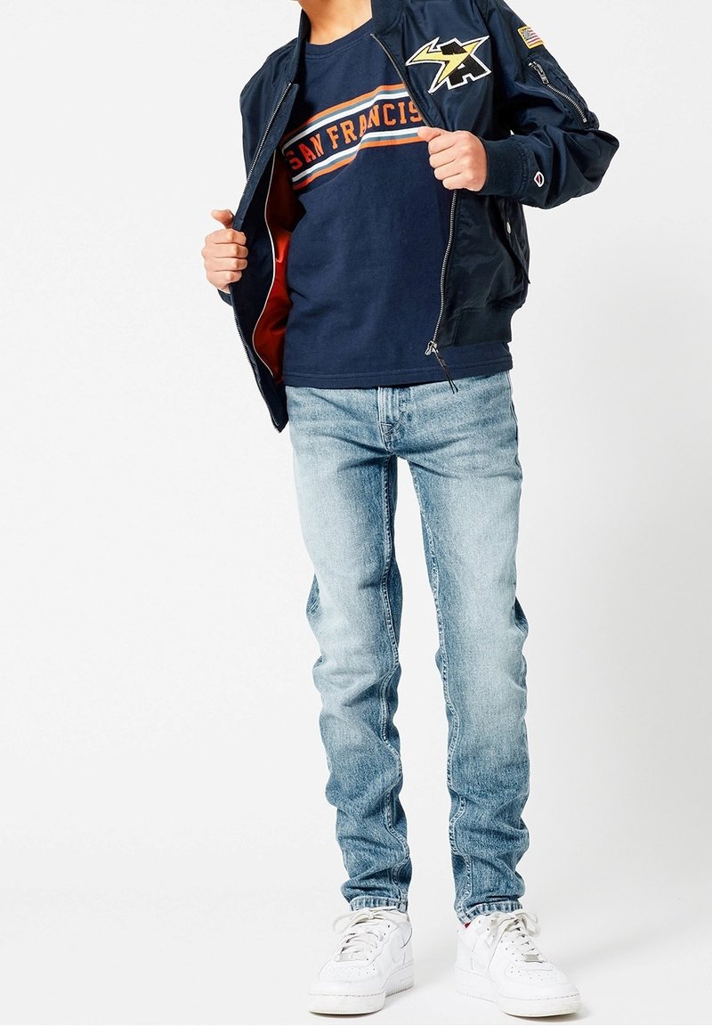 Personne portant une veste bomber bleu marine avec des écussons, un t-shirt bleu marine "San Francisco", un jean bleu clair et des baskets blanches, debout devant un fond blanc.
