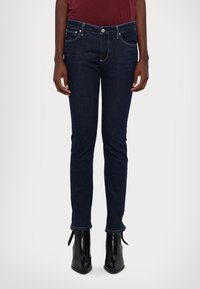 Jeans ajustados azul oscuro de cinco bolsillos, con cremallera y costura en contraste. Combinados con botines negros de tobillo con detalles de atado.