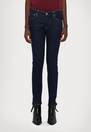 Jeans slim-fit blu scuro con cinque tasche, zip frontale e cucitura a contrasto. Indossati con stivaletti neri alla caviglia con dettagli legati.