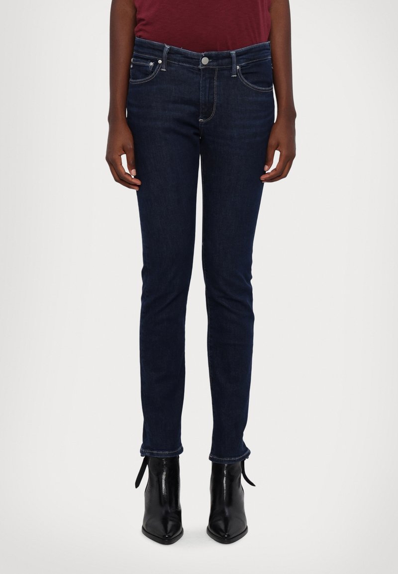 Jeans ajustados azul oscuro de cinco bolsillos, con cremallera y costura en contraste. Combinados con botines negros de tobillo con detalles de atado.