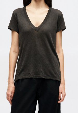 Femme portant un t-shirt gris foncé à manches courtes et col en V, et un pantalon noir, debout devant un fond clair uni.
