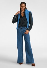 Maglione senza maniche blu indossato sopra una camicia nera con bottoni, abbinato a jeans a gamba larga. Modella con tacchi marroni. Tessuti morbidi.