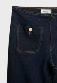 Pantalon en jean bleu foncé avec poche avant boutonnée, bouton en laiton visible, coutures orange et étiquette de taille 38 à la taille.