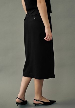 Falda midi negra hecha de una tela suave, con un diseño sencillo, dos bolsillos traseros y un detalle de botón en la cintura. Combinada con tacones negros.