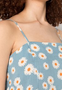 Lichtblauwe jurk met witte madeliefjespatronen en oranje centra. Heeft dunne schouderbanden en een vierkante halslijn. Glad stoftextuur.