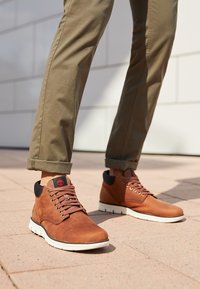 Bottes décontractées à lacets en cuir marron portées avec un pantalon vert olive retroussé, sur un trottoir pavé à l'extérieur.