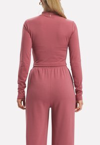 Långärmad jumpsuit i dammig rosa, tillverkad av mjukt tyg, med hög krage och elastisk midja för en bekväm passform.