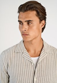 Jasnoszara koszula w paski o miękkiej fakturze, z wyciętym kołnierzem i guzikami. Nosi się ją na białym podkoszulku.