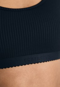 Čierny rebrovaný bralet s výstrihom do tvaru lode a vlnitým lemom. Obsahuje embosované logo "GUESS" na páske. Mäkko textúrovaný materiál.