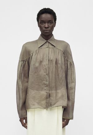 Γυναίκα με κοντά σγουρά μαλλιά που φορά μια φαρδιά, ημιδιαφανή μπλούζα σε χρώμα ταουπέ με μεγάλο γιακά και μακριά μανίκια, συνδυασμένη με ελεφαντόδοντα παντελόνια.