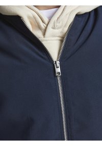 Gros plan d'une fermeture éclair argentée partiellement fermée sur une veste bleu marine portée sur un sweat à capuche beige avec col et cordons visibles.
