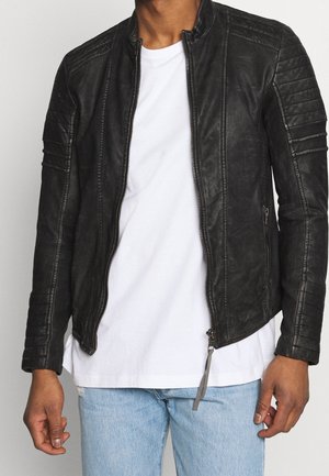 Man wearing a black leather jacket over a white t-shirt and light blue jeans against a plain background.