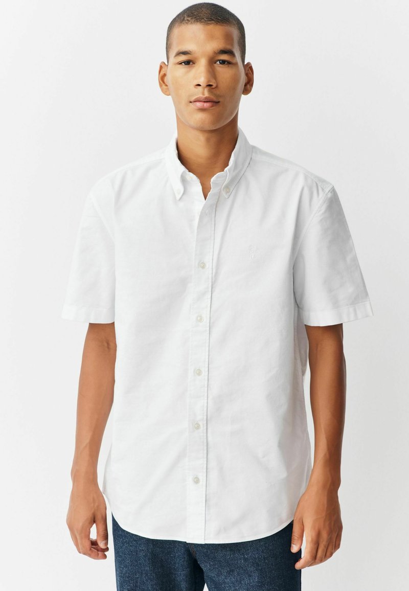 Camisa blanca de manga corta con cuello y botones en la parte delantera, hecha de una tela ligera. Presenta un pequeño logotipo bordado en el pecho.