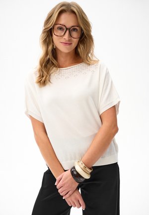 Kobieta o falistych blond włosach, nosząca duże okulary w oprawkach w kolorze żółwia, biały krótki rękaw, czarne spodnie i liczne bransoletki na nadgarstku.