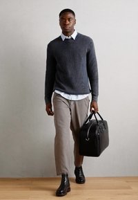 Modellen har på seg en mørkegrå ribbestrikket genser, hvit skjorte med krage, beige bukser og svarte lærdanne støvler, og bærer en svart lærveske.