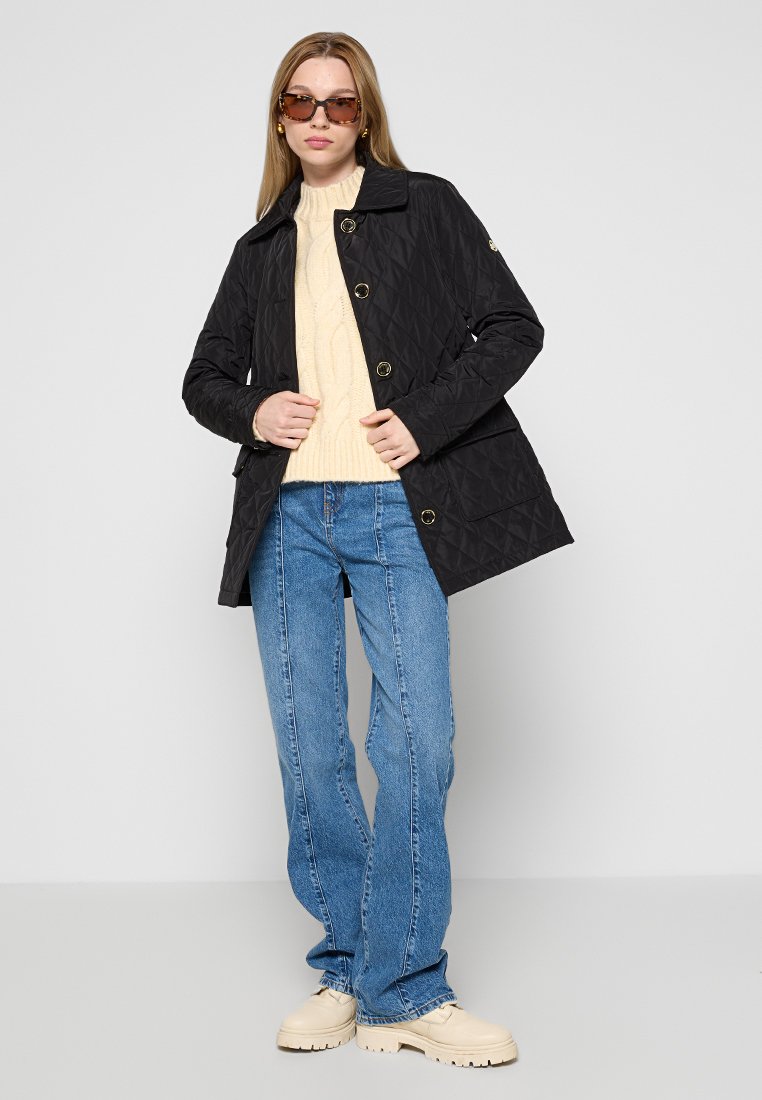 Veste matelassée noire avec col, ornée de boutons dorés. Portée sur un pull crème et associée à un jean bleu et des bottes beiges.