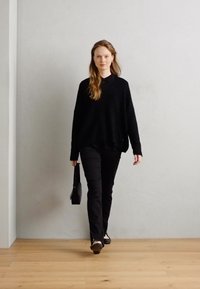 Musthave suur suurusega must kampsun kraega, koos mustade kitsaste pükstega. Kandes musta struktureeritud käsipunutist, kõndides heledal puidust põrandal.