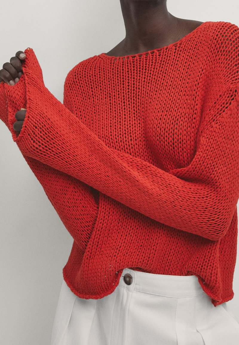 Pull en tricot rouge à coupe ample, col large et manches allongées. Pr�ésente un motif texturé et un ourlet côtelé. Associé à un pantalon blanc.
