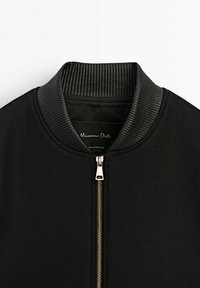 Veste noire avec col en cuir côtelé et fermeture éclair métallique centrale, affichant l’étiquette de la marque "Massimo Dutti" à l’intérieur du col.