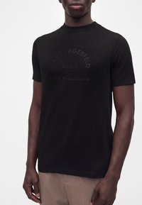 Άνδρας που φοράει μαύρο t-shirt Karl Lagerfeld Paris με ανάγλυφο κείμενο "21 Rue St-Guillaume" και μπεζ παντελόνι, εμφανιζόμενος από τον λαιμό έως τους γοφούς.