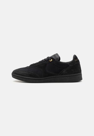Low-profile black sneaker with suede and leather panels, black laces, round toe, and branded heel detail on a white background.