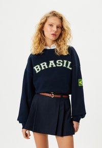 Marineblaues Sweatshirt mit "BRASIL" in grüner Schrift, langen Ärmeln und einem brasilianischen Flaggenpatch. Kombiniert mit einem gefalteten marineblauen Rock und einem braunen Gürtel.