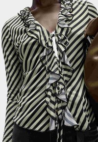 Primo piano di una persona che indossa una blusa a righe diagonali bianche e nere con volant e un papillon al collo, mentre tiene una borsa marrone.