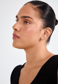 Brinco de argola prateada numa modelo com um coque elegante, apresentando uma textura suave e um acabamento brilhante, contra um fundo neutro.