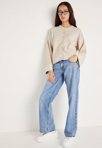 Μπεζ καρουλένιο cardigan με φαρδιά μανίκια συνδυασμένο με ανοιχτό μπλε τζιν ίσιου κοψίματος και λευκά αθλητικά παπούτσια σε ουδέτερο φόντο.