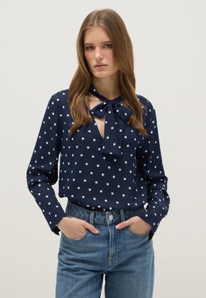 Jeune femme portant un chemisier à manches longues bleu marine à pois blancs avec un nœud noué au niveau du col, associé à un jean bleu.