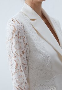 Biały żakiet z koronkowymi rękawami w kwiatowe wzory. Gładki materiał z kołnierzem szalowym i jednorzędowym zapięciem.